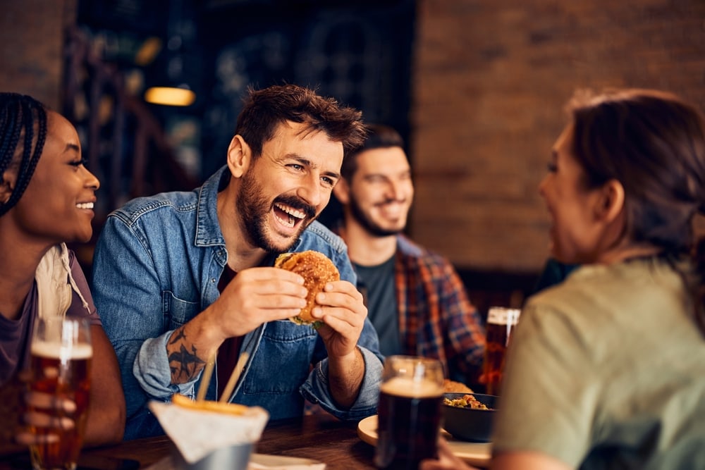 Cheerful man eating burger and having fun while gathering with friends in a bar. Best burger cities in american